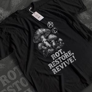 Puede incluir: Camiseta negra con una ilustración detallada de setas y el texto "ROT, RESTORE, REVIVE!". El diseño incluye los símbolos "A" y "E". Una etiqueta blanca está sujeta a la camiseta.