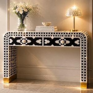 May include: A black and white patterned console table with two drawers, gold accents, and a curved design. The table is decorated with a vase of white flowers, books, and a decorative lamp.