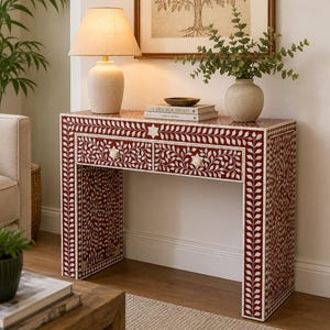 May include: A rectangular console table featuring a deep red surface with intricate white floral and leaf inlay patterns. The table has two drawers with star-shaped knobs, a flat top surface, and four sturdy legs, all covered in a consistent botanical design.