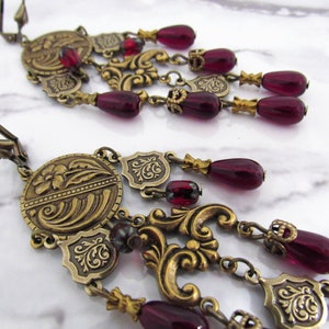 May include: A pair of gold tone dangle earrings with burgundy glass beads and ornate metal details.
