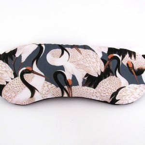 May include: A sleep mask featuring a pattern of white cranes with black and orange accents against a dark teal background. The mask has a curved shape with rounded edges and small fabric loops on each side.