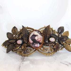 May include: An antique-style hair barrette with a central oval portrait, surrounded by ornate bronze-toned metalwork and detailed leaf accents. The barrette features two iridescent, oval-shaped stones and is designed for decorative hair styling.