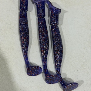 May include: Three purple soft plastic fishing lures with a paddle tail design. Each lure features a translucent deep purple body embedded with small, reflective red and silver glitter flakes throughout the entire length of the bait.