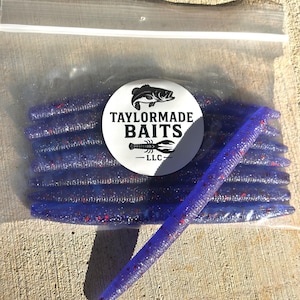 May include: A clear plastic bag containing several purple fishing lures with red flecks. The lures are cylindrical and tapered. A white circular label with the text "TAYLORMADE BAITS" and a fish graphic is visible.