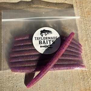 May include: A clear plastic bag filled with purple fishing lures. The lures are cylindrical with a textured surface and glitter accents. The bag has a white circular label with the text "TAYLORMADE BAITS" and a fish graphic.