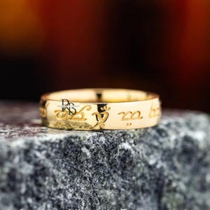 Puede incluir: Un anillo dorado con escritura grabada. El anillo está sobre un fondo borroso y descansa sobre una superficie gris texturizada. El diseño del anillo presenta letras estilizadas e intrincadas alrededor de su banda.