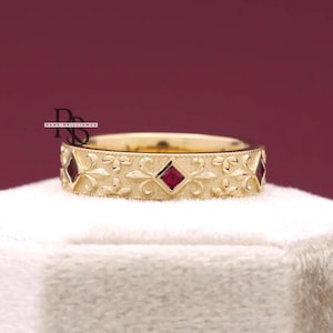 May include: A gold-colored ring with a repeating floral and diamond pattern. The ring features small, square, red gemstones set within the diamond shapes. The ring is displayed on a white surface against a dark red background.