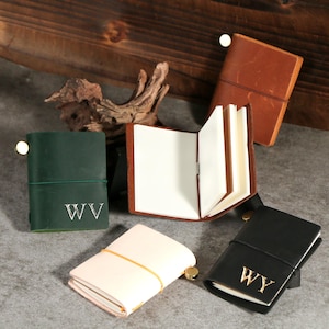 May include: A selection of small leather journals in shades of dark green, cream, black, and brown. Each journal is secured with an elastic band and features a gold-coloured button closure. The journals are personalised with gold initial lettering.