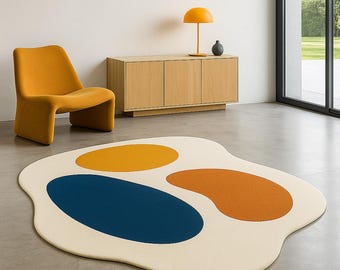 May include: A modern, abstract area rug with a cream-colored base and three large, colorful shapes: a blue oval, a yellow circle, and an orange kidney bean shape. The rug is on a concrete floor, with a mustard-colored chair and wooden cabinet in the background.