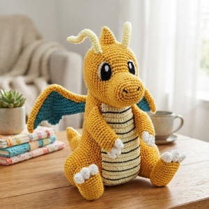 Puede incluir: Un peluche de dragón tejido a ganchillo con cuerpo amarillo dorado, alas color verde azulado y una barriga color crema con rayas horizontales negras. La criatura tiene dos antenas, garras blancas en manos y pies, y grandes ojos negros y blancos.