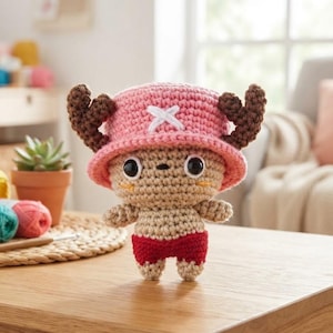 Puede incluir: Una figura amigurumi de ganchillo con cuerpo color canela, pantalones cortos rojos y un sombrero rosa con astas marrones y un emblema de cruz blanca. El personaje tiene grandes ojos negros, una nariz pequeña y dos líneas naranjas en las mejillas.