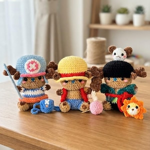 Op de afbeelding: Drie gehaakte knuffelfiguren geïnspireerd op anime-personages. De linkerfiguur draagt een blauwe hoed met een roze kruis, een gestreept shirt en een blauwe tas. De middelste figuur draagt een gele strooien hoed en een rood vest. De rechterfiguur draagt een zwarte hoed met een klein wit wezen erop en een groen gewaad.