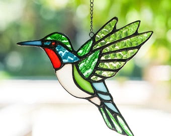 May include: A stained glass hummingbird suncatcher featuring vibrant green, red, blue, and white glass pieces held together by black metal solder lines. The bird is depicted in flight with wings extended and is suspended by a thin metal chain.