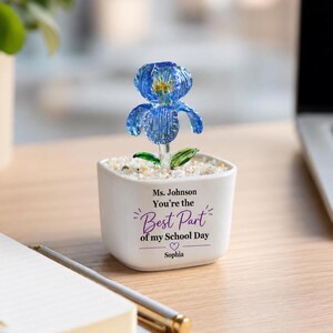 May include: A blue glass iris flower with green leaves sits in a white square ceramic planter filled with small white stones. The planter features black and purple text that reads: Ms. Johnson You're the Best Part of my School Day Sophia.