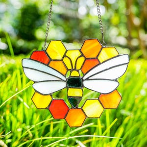 May include: A stained glass bee suncatcher with a honeycomb design. The bee has white wings, a yellow and black body, and is surrounded by yellow, orange, and red hexagonal glass pieces. It hangs from a metal chain against a blurred green background.