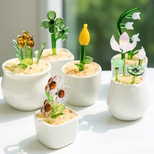 Könnte beinhalten: Eine Sammlung von Glasblumenskulpturen in kleinen weißen Keramiktöpfen. Die Blumen umfassen Tulpen, Kleeblätter und Gänseblümchen mit grünen Stielen und bunten Blütenblättern. Die Töpfe sind mit einem hellfarbigen, sandigen Material gefüllt.