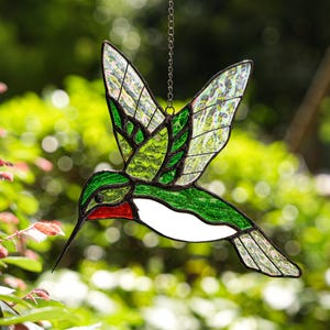 Puede incluir: Atrapasoles de colibrí de vidrieras con detalles verdes, rojos y blancos. Las alas son una mezcla de vidrio verde y transparente, y el pájaro cuelga de una cadena plateada. El fondo es una vista borrosa de follaje verde.