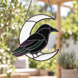 May include: A stained glass suncatcher featuring a black crow perched on a white crescent moon. The crow has iridescent purple and green wing accents, and the entire piece is framed in dark metal solder, suspended by a thin metal chain.