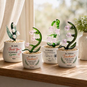 May include: Four white ceramic pots containing glass lily of the valley flowers with green stems and white or pink blossoms. Each pot features personalized text and decorative graphics, including phrases like Thank You for helping me Grow and Teachers change the world one student at a time.