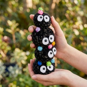 May include: A stack of four black plush toy figures with large white eyes and colorful pom-pom accents. The figures are held in a person's hands against a blurred green background.