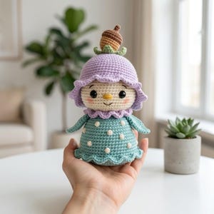 May include: A crochet doll featuring a cream-colored face with black eyes and pink cheeks, wearing a lavender hat topped with a brown acorn. The doll has a teal dress with white polka dots, a ruffled collar, and a matching ruffled hemline.