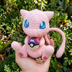 May include: A handmade pink crochet Mew plushie with blue eyes and a long tail. It holds a purple and pink Poké Ball. The plushie has a small wooden tag that says "HANDMADE".