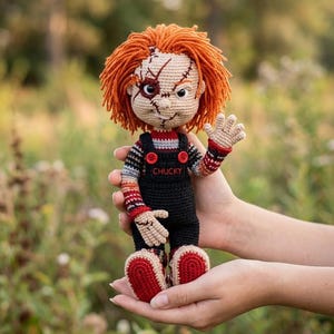May include: A handmade crochet doll of Chucky, the horror movie character. The doll has orange hair, a stitched scar on its face, and is wearing black overalls with red buttons. The doll's hands and feet are beige with red accents. The word "CHUCKY" is visible.