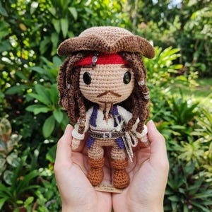 May include: A handmade crochet doll of a pirate character, featuring a brown tricorn hat, red bandana, and long brown dreadlocks. The doll wears a white shirt, purple vest, and brown boots. It has a small mustache and a toy sword. The doll is held in someone's hands.
