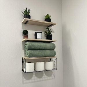 May include: A bathroom shelf arrangement featuring three wooden shelves with black metal brackets. The top shelves hold small potted plants. The middle shelf displays a candle and two green towels. The bottom shelf has three rolls of toilet paper.