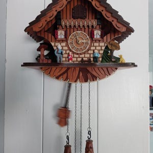 May include: A wooden cuckoo clock with a detailed, multi-tiered design. The clock features a pitched roof, a round clock face with Roman numerals, and decorative figurines on a shelf. Two pinecone-shaped weights hang below, along with a pendulum.
