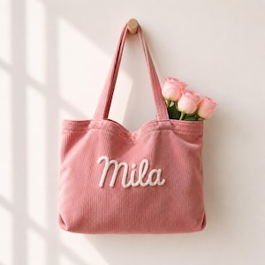 Könnte beinhalten: Eine altrosa Tragetasche aus Kord mit zwei Schulterriemen und dem in weißer Schreibschrift aufgestickten Namen Mila auf der Vorderseite. Drei hellrosa Rosenblüten stecken in der Tasche und ragen über den oberen Rand hinaus.