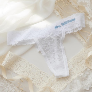 Pode incluir: Cueca tanga de renda branca com o texto bordado "Mrs. Williams" em azul claro. A lingerie delicada apresenta um pequeno laço e bordas onduladas. O fundo inclui tecido branco transparente e acabamento em renda.