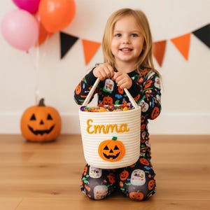 May include: A white rope basket with the name "Emma" embroidered in yellow, and a felt pumpkin applique. The basket is filled with candy and held by a child wearing Halloween pajamas. Orange and pink balloons are in the background.