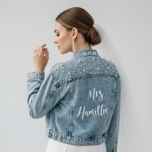 Könnte beinhalten: Hellblaue Jeansjacke mit Perlenbesatz und dem Text "12.11.25" auf dem oberen Rücken. Die Jacke trägt auch den Namen "Mrs. Hamilton" in weißer Schrift auf dem unteren Rücken. Eine Brautjacke.