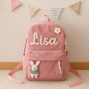Puede incluir: Una mochila rosa de pana con el nombre "Lisa" en escritura blanca. La mochila presenta un bolsillo frontal, una flor blanca y un pequeño conejito de peluche. Las correas rosas y un asa superior completan el diseño. Banderines decorativos cuelgan en el fondo.