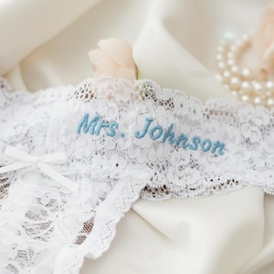 May include: White lace garter with the embroidered text "Mrs. Johnson" in light blue. The garter features a delicate bow and is displayed on a white satin surface, with a pearl necklace and a rose in the background.