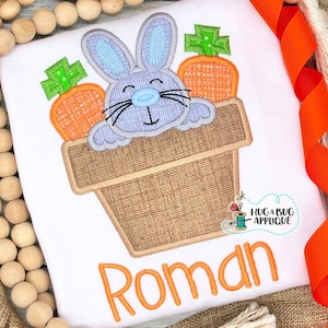 Puede incluir: Un conejito azul claro asomándose a una maceta marrón con dos zanahorias naranjas y hojas verdes. El texto "Roman" está bordado debajo de la maceta.