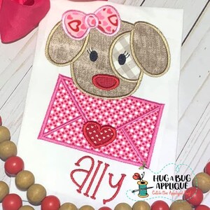 Può includere: Applicazione ricamata con un cane marrone che tiene una busta rosa e bianca con un cuore rosso. Il nome "ally" è ricamato sotto il cane. Il design è su una maglietta bianca.