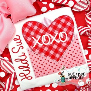Può includere: Una maglietta bianca con un motivo a cuore a quadri rossi e rosa e le lettere ricamate "XOXO". La maglietta presenta anche il nome "Libbi Sue" in corsivo rosso. Il design è circondato da fiocchi e nastri rossi e rosa.