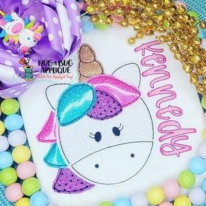 May include: A white shirt with a pink unicorn applique featuring a blue and purple mane and a gold horn. The shirt also has the name "Kennedy" embroidered in pink.
