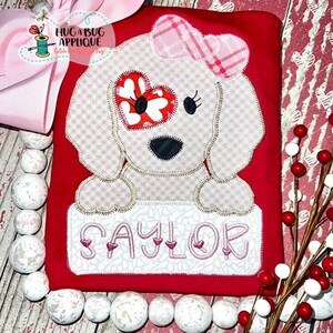 May include: A white felt applique of a dog with a pink bow and a heart-shaped eye. The dog is holding a banner with the name "Saylor" embroidered in pink.