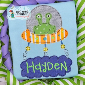 May include: A light blue t-shirt with an embroidered design of a green alien in a spaceship with yellow lights and a cloud with the name Hayden in green letters.