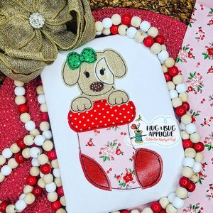 May include: A white t-shirt with an embroidered design of a brown dog peeking out of a pink and red stocking with white polka dots. The text "Hug a Bug Applique" is embroidered on the shirt.