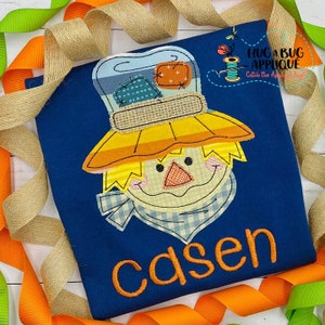 Op de afbeelding: Een blauw T-shirt met een kleurrijke patchwork-vogelverschrikker applicatie en de naam "Caden" geborduurd in oranje draad.