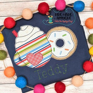 Könnte beinhalten: Ein blaues T-Shirt mit einem gestickten Design einer Kaffeetasse mit Schlagsahne und einem Donut. Der Text "Teddy" ist unter dem Design gestickt.