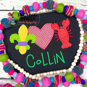 Puede incluir: Aplicación negra con una flor de lis, un corazón de cuadros y un cangrejo rojo. El nombre "Collin" está bordado en verde. La aplicación está rodeada por una colorida guirnalda de cuentas. El texto en la parte superior izquierda dice "HUGA BUG APPLIQUE Catch the Applique Bug!"