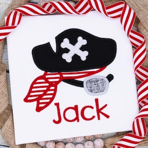 May include: White t-shirt with an embroidered black pirate hat, red and white striped bandana, and a black eyepatch. The name "Jack" is embroidered in red below the pirate design.