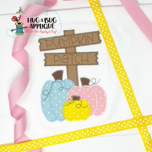 May include: A white fabric with a brown wooden sign that says "Pumpkin Patch". There are three pumpkins below the sign, one blue, one pink, and one yellow.