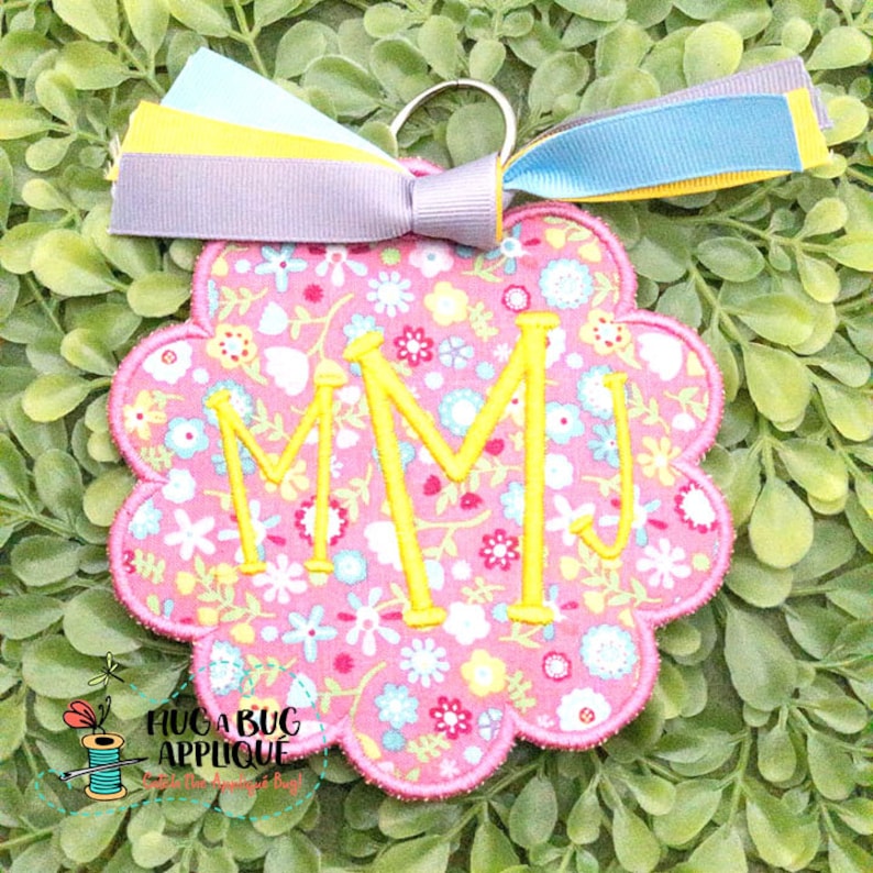 May include: A pink and yellow floral fabric key chain with a scalloped edge and the letters "MM" embroidered in yellow. The key chain has a silver key ring and a multi-colored ribbon attached.