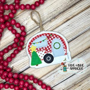 May include: A red and white camper with a Christmas tree and lights, a wreath on the door, and a dragonfly on the side. The camper is a felt applique ornament with a loop for hanging.